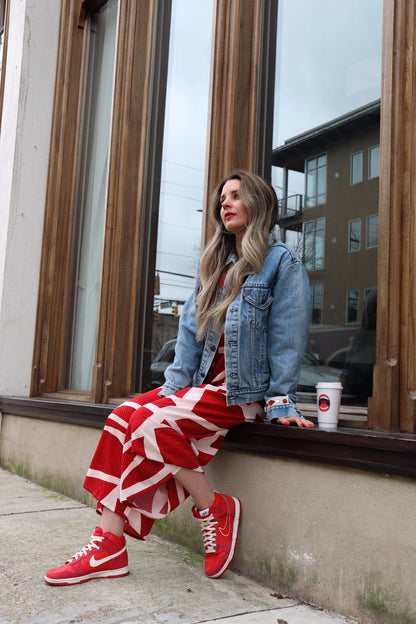  hailey is a white woman with long medium brown wavy hair. she is wearing a kaarem red and white printed overall-style jumpsuit with a one of a kind denim jacked. she has red nike sneakers and red lipstick to match.