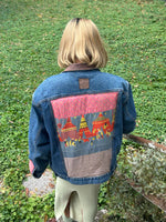 mary has short blonde hair and she walks in her garden wearing an upcycled denim jacket with a quilted center back panel of a circus over a fitted pale yellow dress