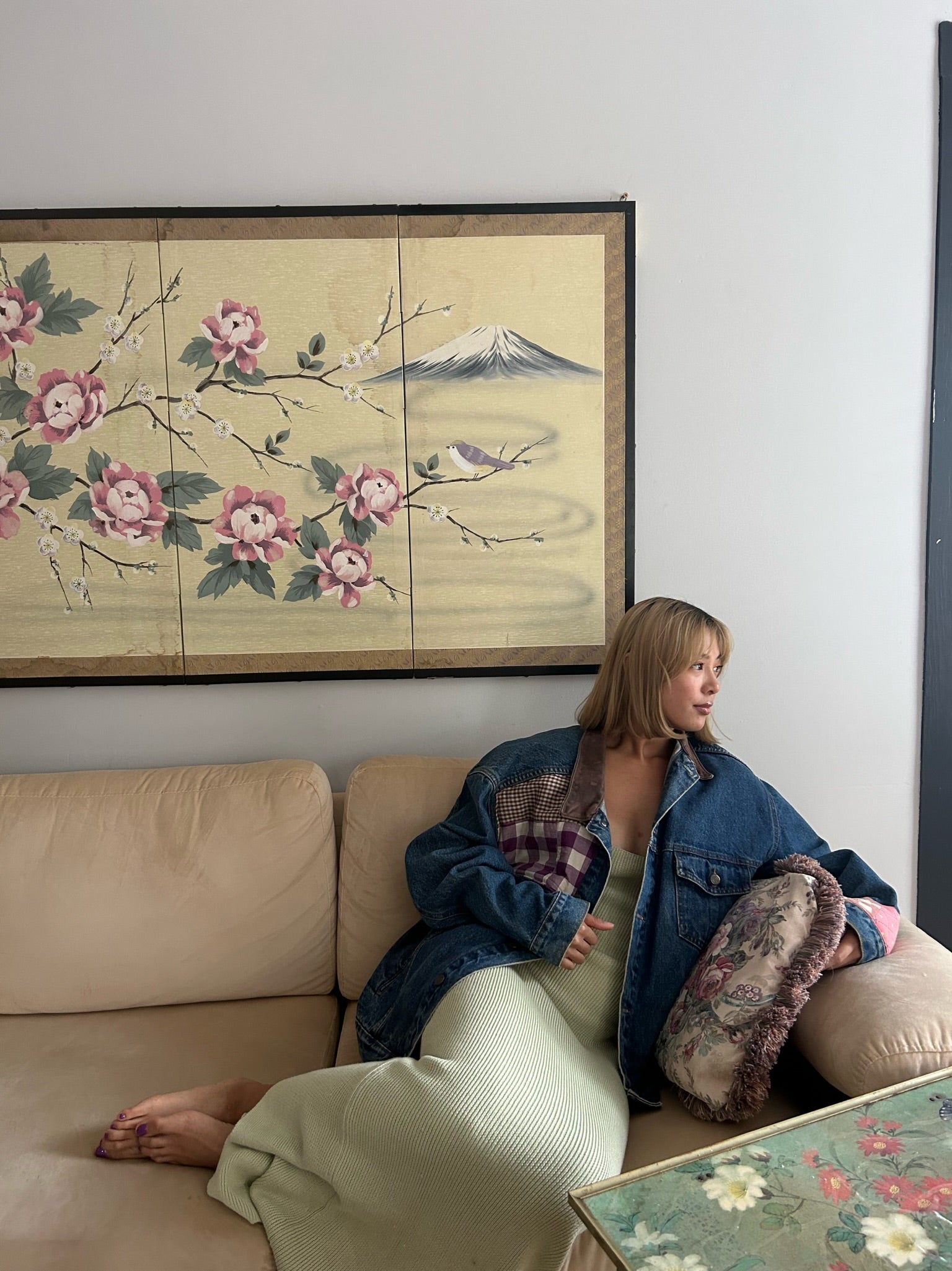mary has short blonde hair sitting on a couch looking over her shoulder. she is wearing an upcycled denim jacket with a quilted center back panel of a circus over a fitted pale yellow dress