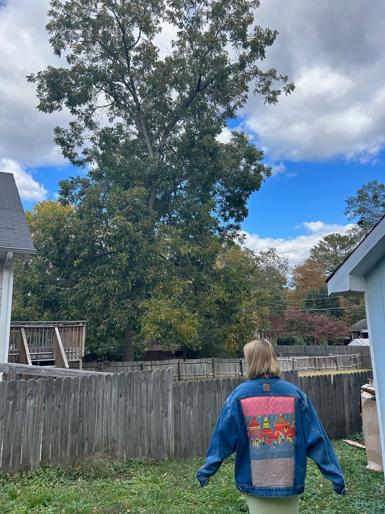 mary has short blonde hair and she walks in her garden wearing an upcycled denim jacket with a quilted center back panel of a circus over a fitted pale yellow dress