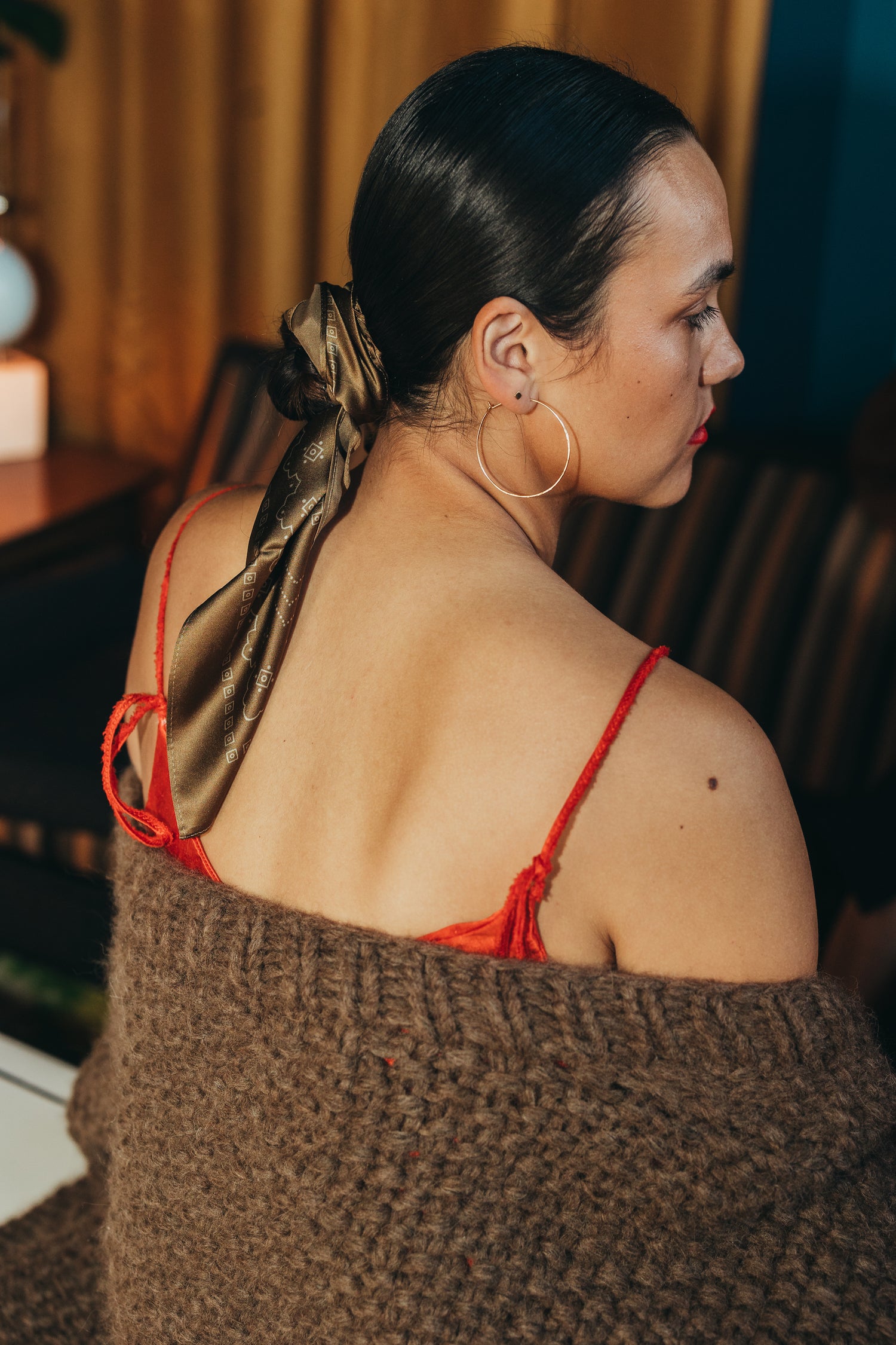 detail shot of phoebe, a latina woman, wearing the danielle dean little red dress under an oversized brown lulua cardigan by nia thomas. she has a scarf by manner market wrapped around her low bun. she is wearing large gold hoops by ember&amp;onyx.