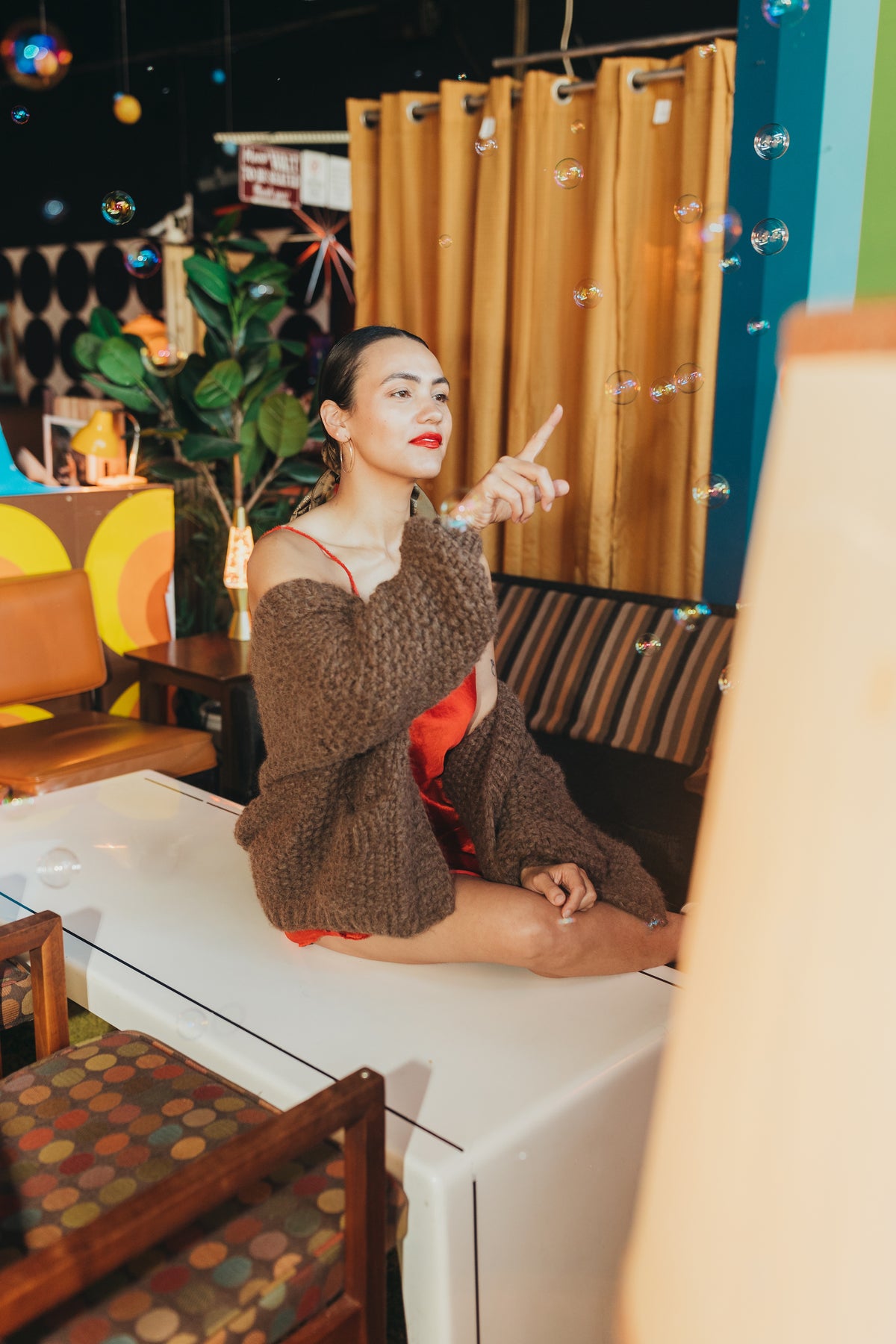 phoebe is wearing the danielle dean little red dress with the nia thomas lulua cardigan. she is looking away from the camera, and the cardigan falls off her shoulders. her hair is pulled back low in a bun wrapped in a manner market silk scarf. she is trying to pop a bubble.