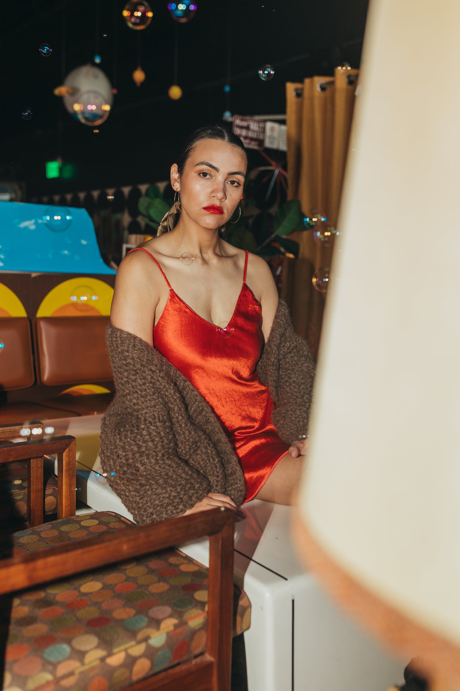 phoebe is a latina woman wearing the danielle dean little red dress under an oversized brown lulua cardigan by nia thomas. she is sitting on a white table with bubbles and chaos around her.
