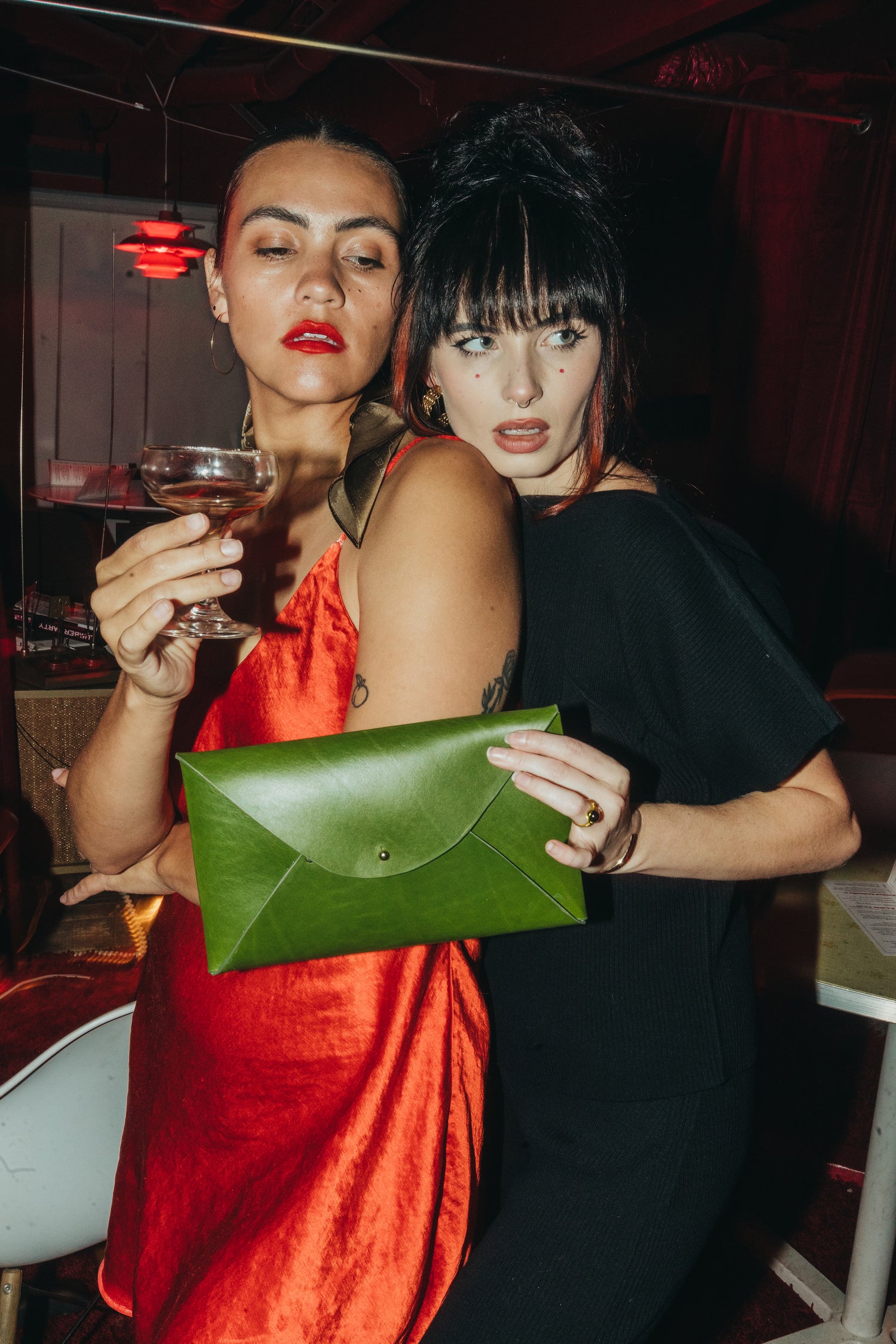 phoebe is a latina woman wearing the danielle dean little red dress drinking a martini while moe, a white woman wearing the nia thomas mujer + hombre set, holds a green envelope clutch by elidia the label.