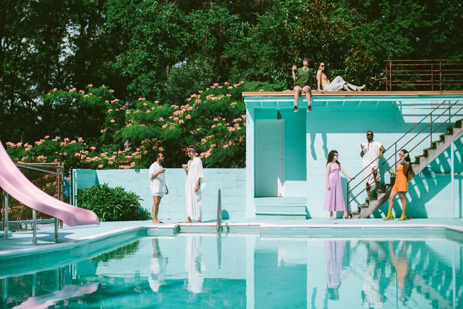 a slim aarons-inspired photoshoot for pride 2020. 7 people stand on an aqua blue pool deck á la palm springs 1960. a pink slide peeks in from the left.