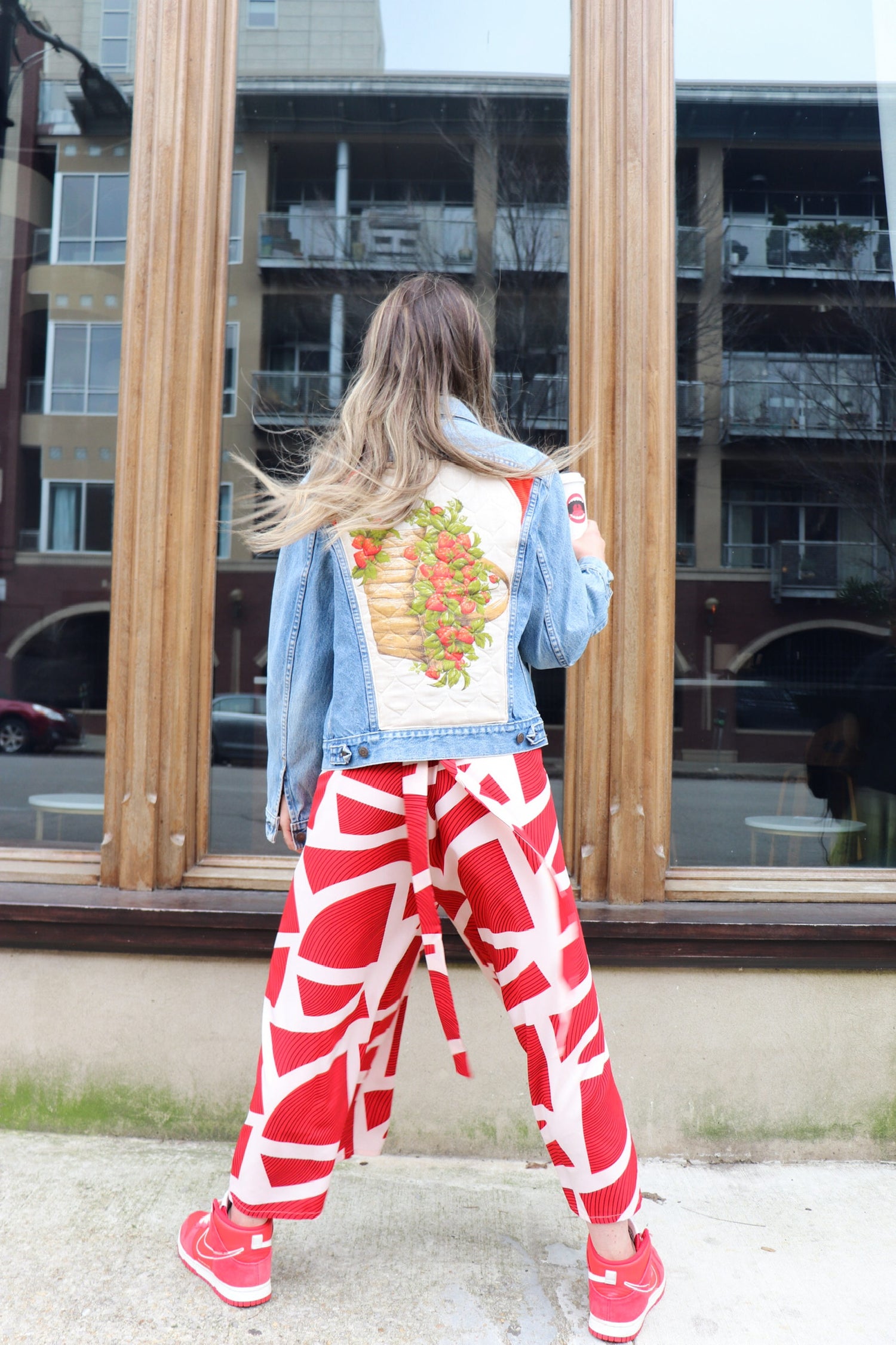  hailey is a white woman with long medium brown wavy hair. she is standing with her back to us wearing a kaarem red and white printed overall-style jumpsuit with a one of a kind denim jacket with a quilted strawberry patch in the back center panel.. she has red nike sneakers to match.