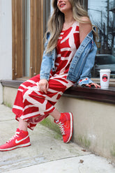 hailey is a white woman with long medium brown wavy hair. she is wearing a kaarem red and white printed overall-style jumpsuit with a one of a kind denim jacked. she has red nike sneakers and red lipstick to match.