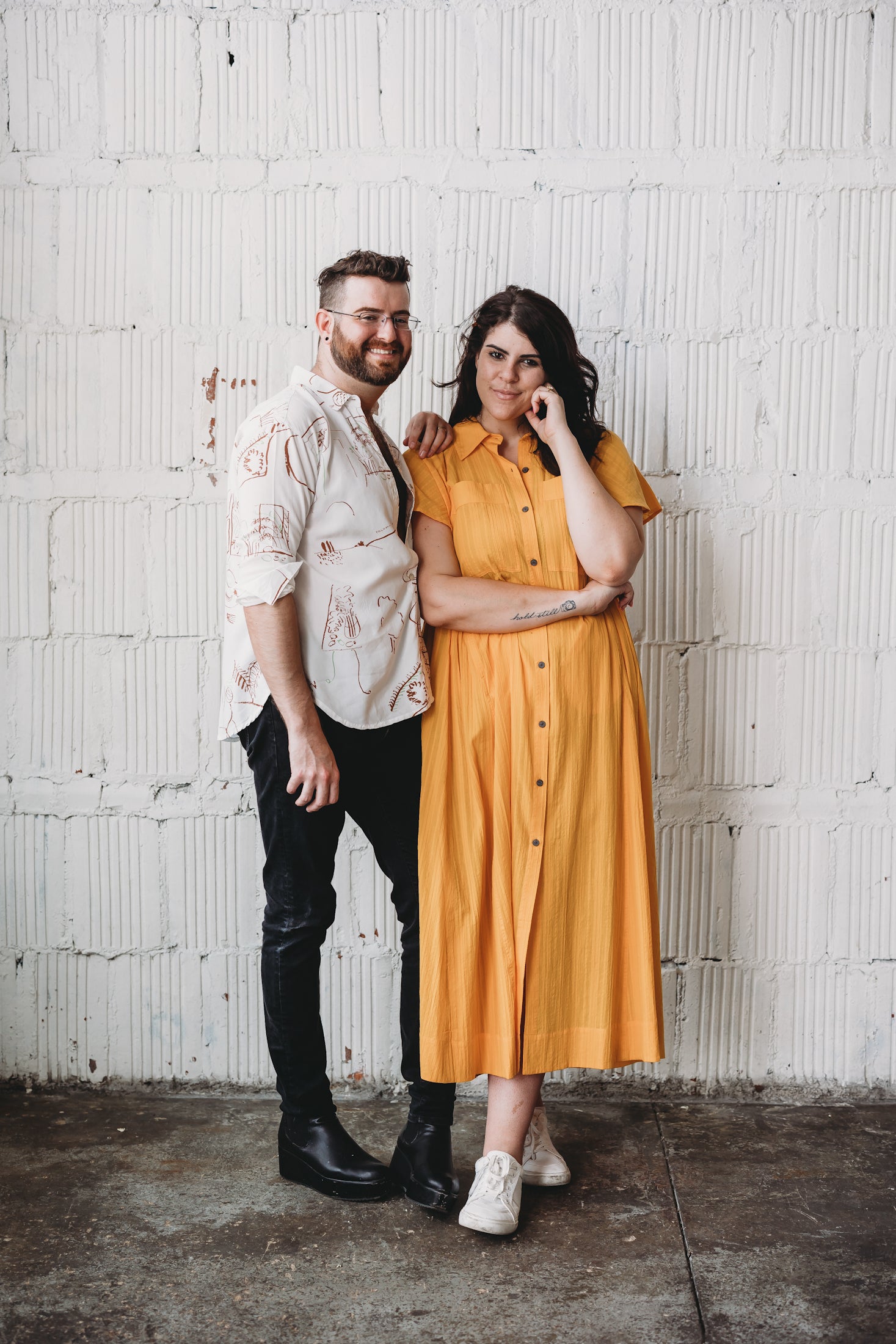 robbie stands on the left facing inwards. he is wearing glasses and has on a white button down with line drawings on it paired with black jeans. His hand rests on Mary's shoulder. Mary is wearing a long, tangerine button-up shirtdress with white sneakers