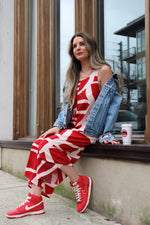 hailey is a white woman with long medium brown wavy hair. she is wearing a kaarem red and white printed overall-style jumpsuit with a one of a kind denim jacked. she has red nike sneakers and red lipstick to match.