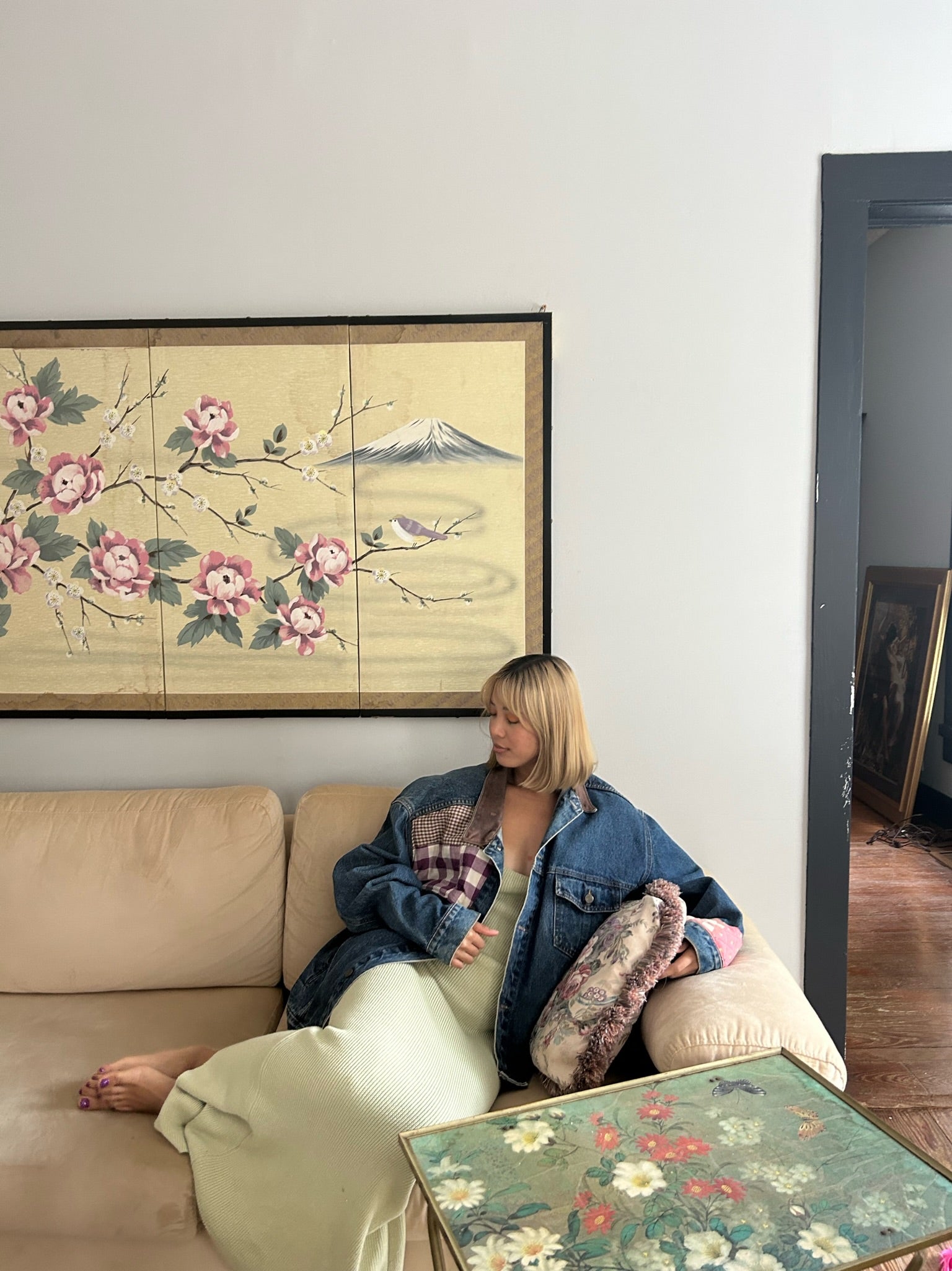 mary has short blonde hair sitting on a couch looking over her shoulder. she is wearing an upcycled denim jacket with a quilted center back panel of a circus over a fitted pale yellow dress