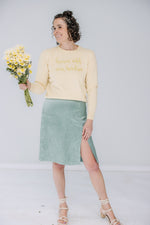 lacey is a white woman with dark, shoulder-length curly hair. she's holding a bouquet wearing a yellow lingua franca sweater that says "bans off our bodies" and the danielle dean mamie skirt in a mermaid green.