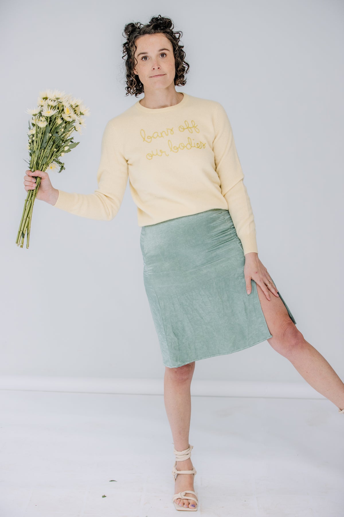 lacey is a white woman with dark, shoulder-length curly hair. she's holding a bouquet wearing a yellow lingua franca sweater that says "bans off our bodies" and the danielle dean mamie skirt in a mermaid green.