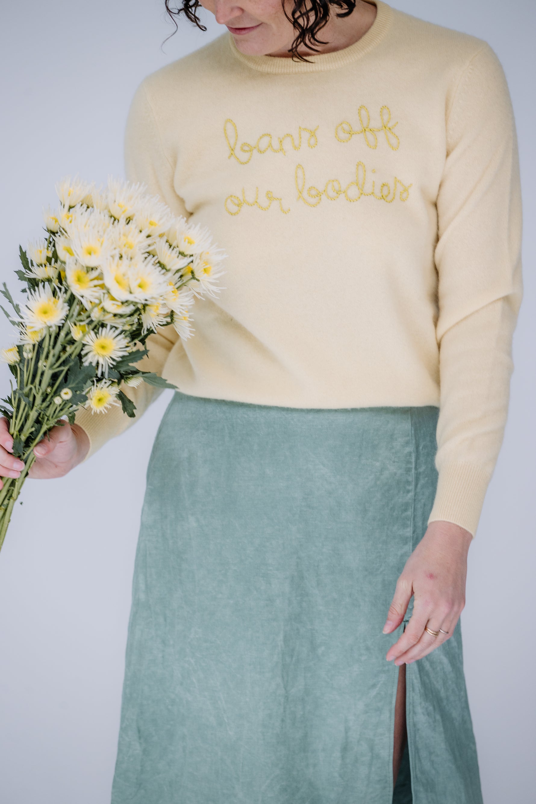 lacey is a white woman with dark, shoulder-length curly hair. she's holding a bouquet wearing a yellow lingua franca sweater that says "bans off our bodies" and the danielle dean mamie skirt in a mermaid green.