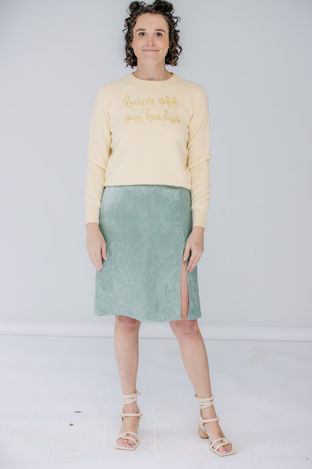 lacey is a white woman with dark, shoulder-length curly hair. she's holding a bouquet wearing a yellow lingua franca sweater that says "bans off our bodies" and the danielle dean mamie skirt in a mermaid green.