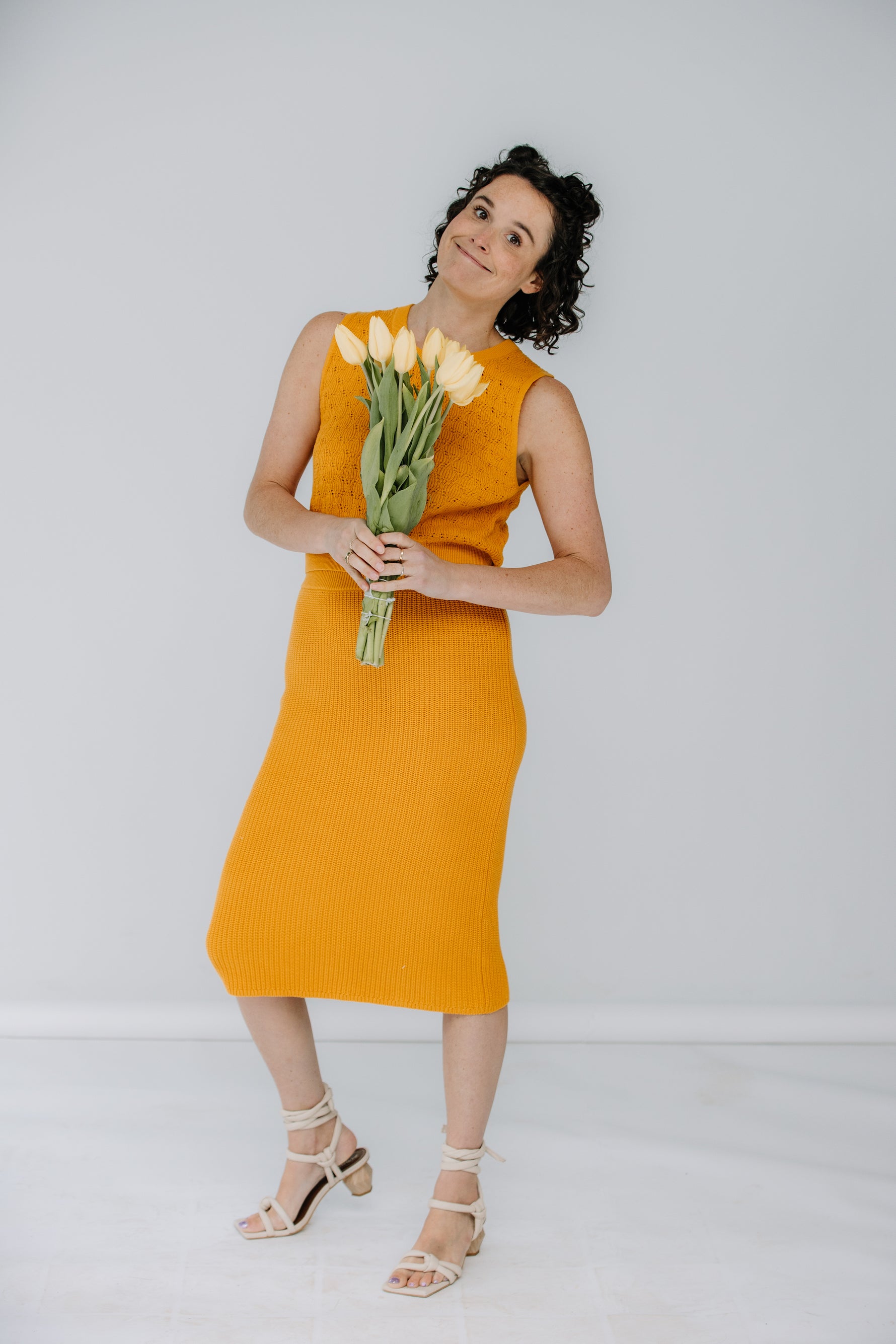 lacey wears the pointelle top and ribbed midi skirt in marigold by mr mittens. she is holding yellow tulips and has aloha creative ivory heels on.