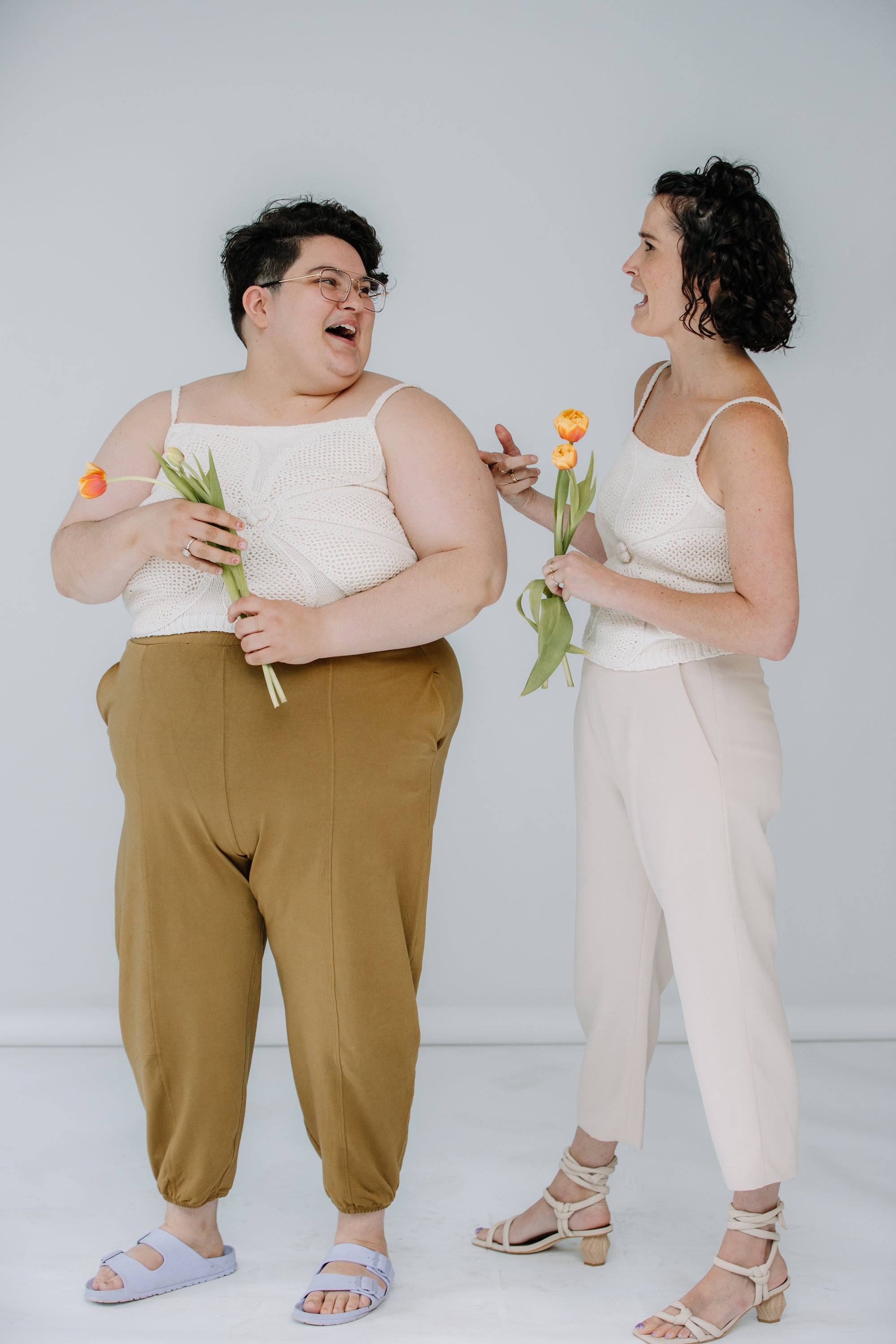 jules and lacey model the lingua franca crochet flower tank in ivory. jules is wearing theirs with mustard day lantern pants, and lacey has on kaarem sua trousers. they are both wearing sandals and talking.