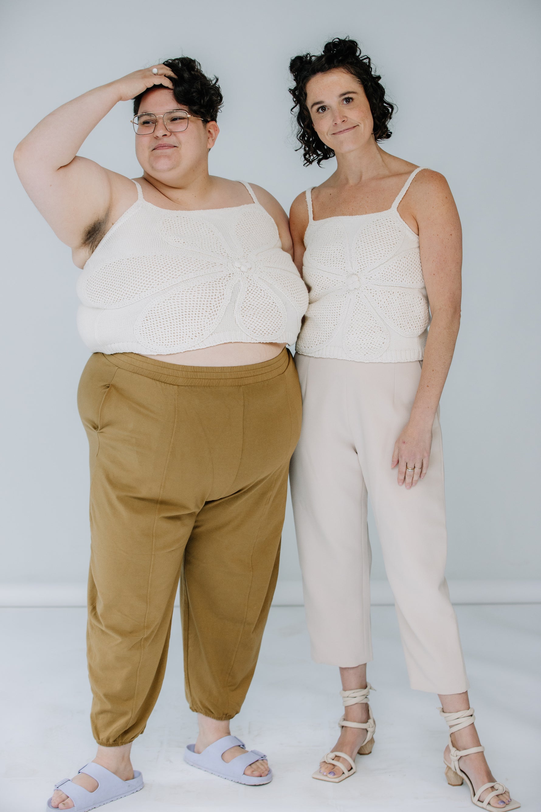 jules and lacey model the lingua franca crochet flower tank in ivory. jules is wearing theirs with mustard day lantern pants, and lacey has on kaarem sua trousers. they are both wearing sandals and soft smiling.