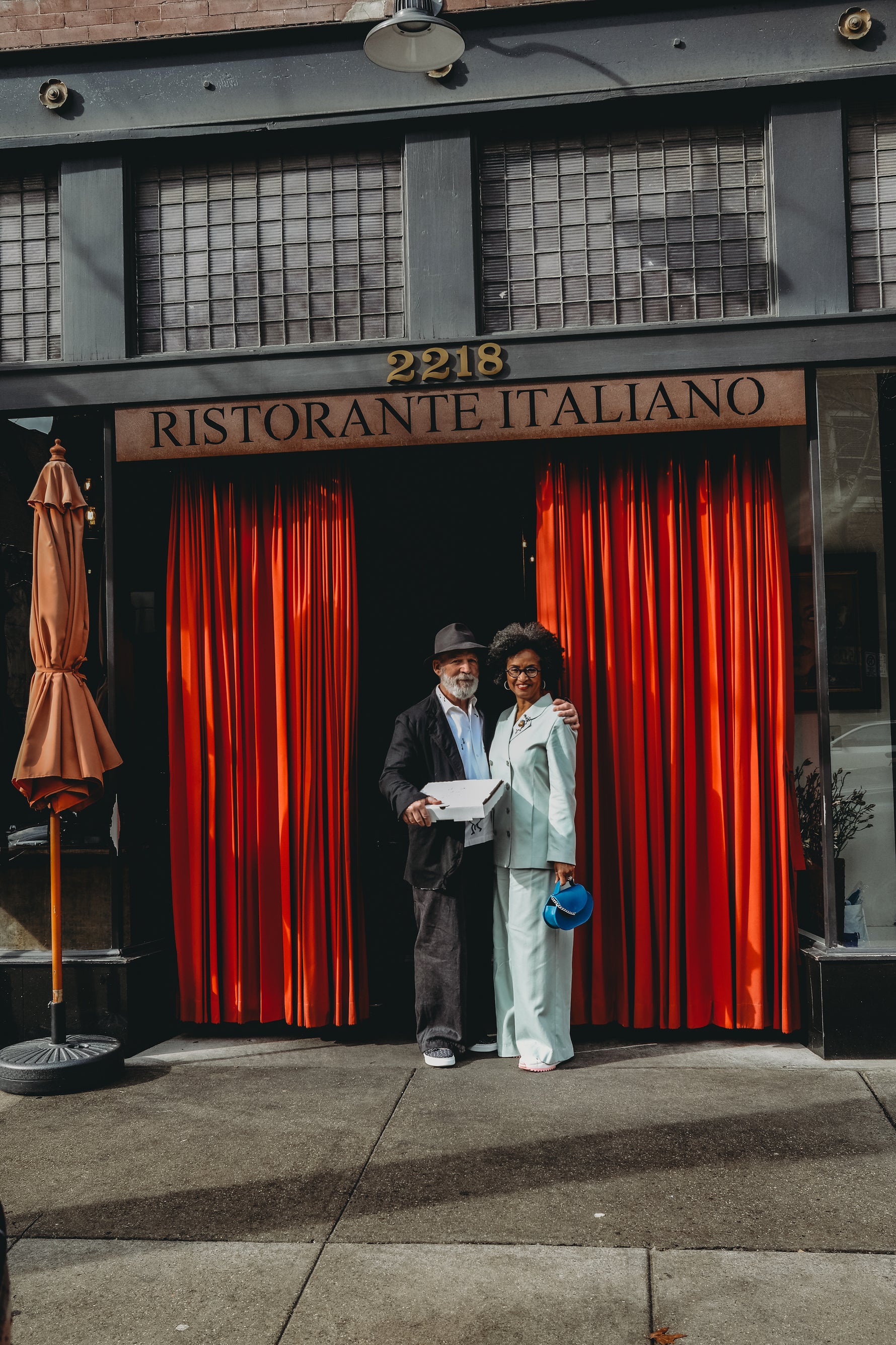 val and steve stand smiling in front of red drapes that lead into Le Fresca. steve is wearing a black bowler cap and black suit while valerie wears the dylan pants + freya blazer from basic.