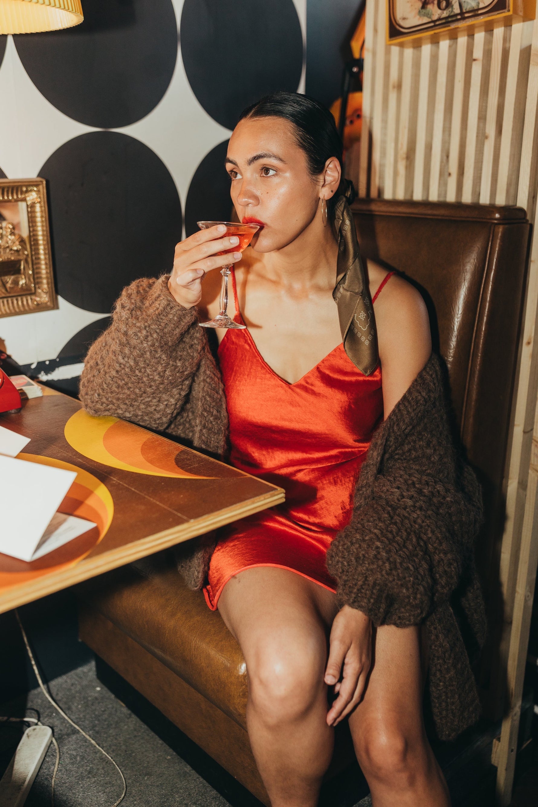 phoebe is a latina woman wearing the danielle dean little red dress under an oversized brown lulua cardigan by nia thomas. she is sitting in a bar booth drinking a martini.