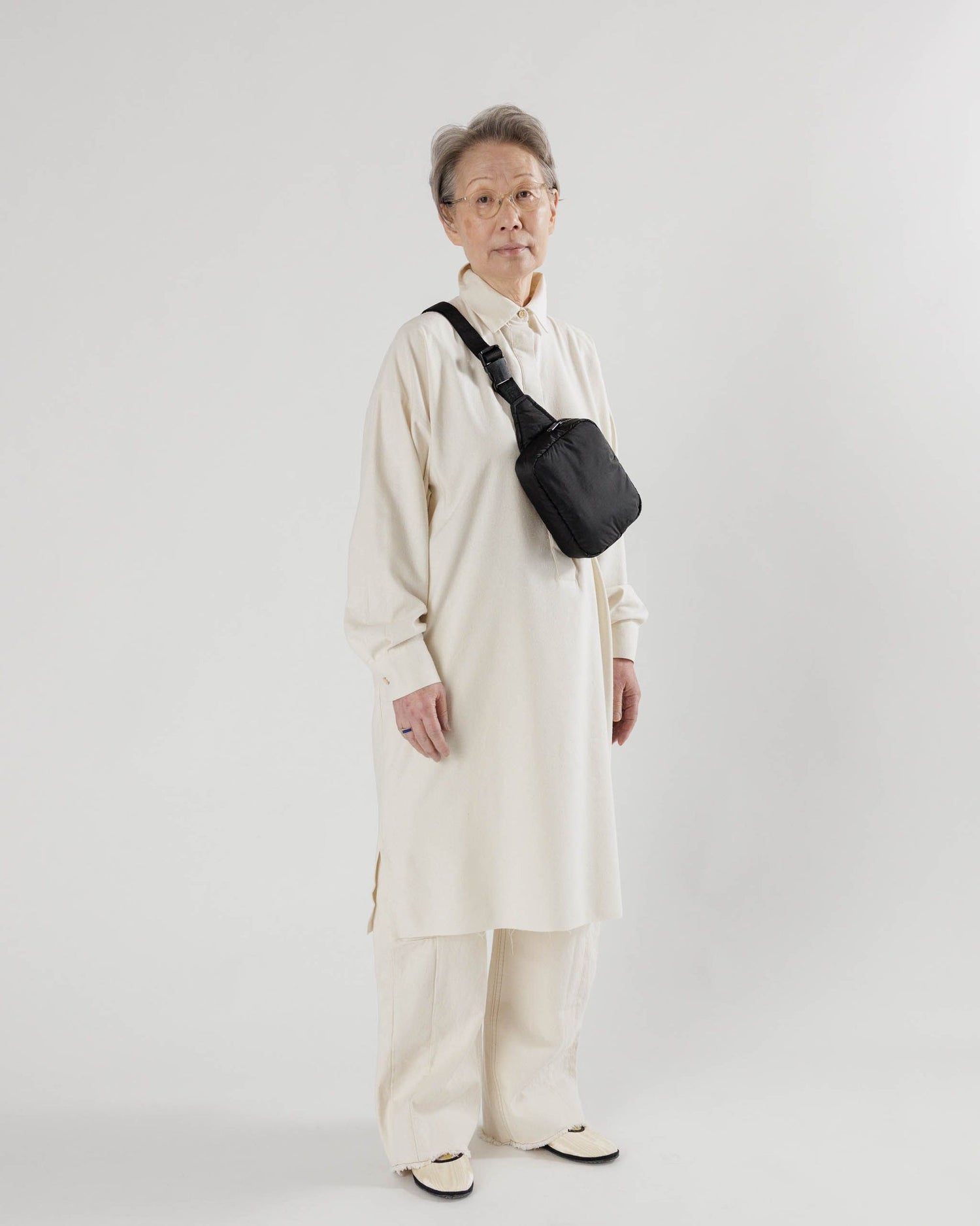 an older woman with short gray hair wears a long white tunic over white sweats paired under a black baggu puffy fanny pack.