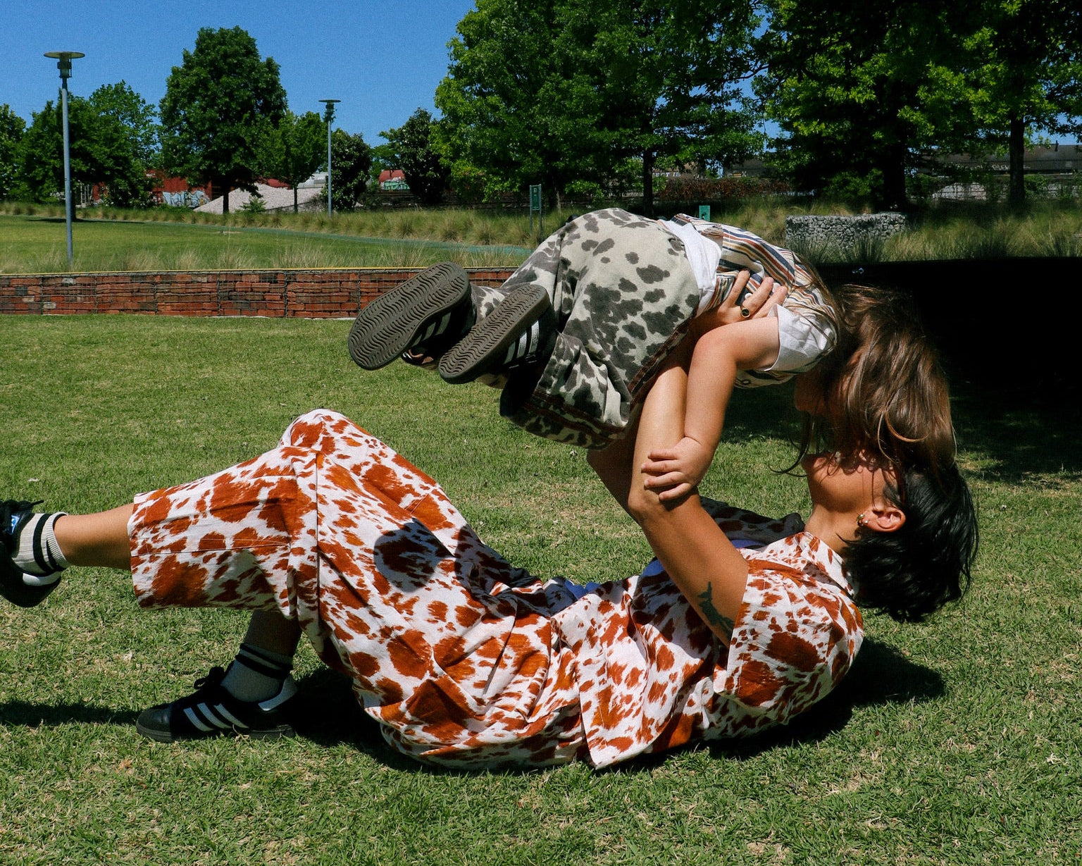 our friend and her child play together on a grassy park lawn under a blue sky with trees in the background. The woman is wearing a loose white dress with a brown and orange abstract pattern, and the child is wearing a gray and white patterned top and shorts.