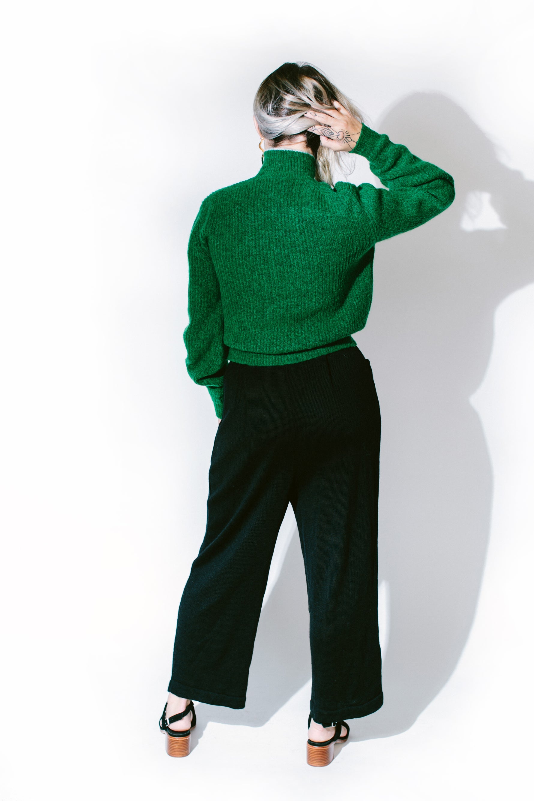 lemon stands with her back to the camera wearing a paloma wool sweater and été noir high-waisted alpaca trousers in front of a white background - basic. 